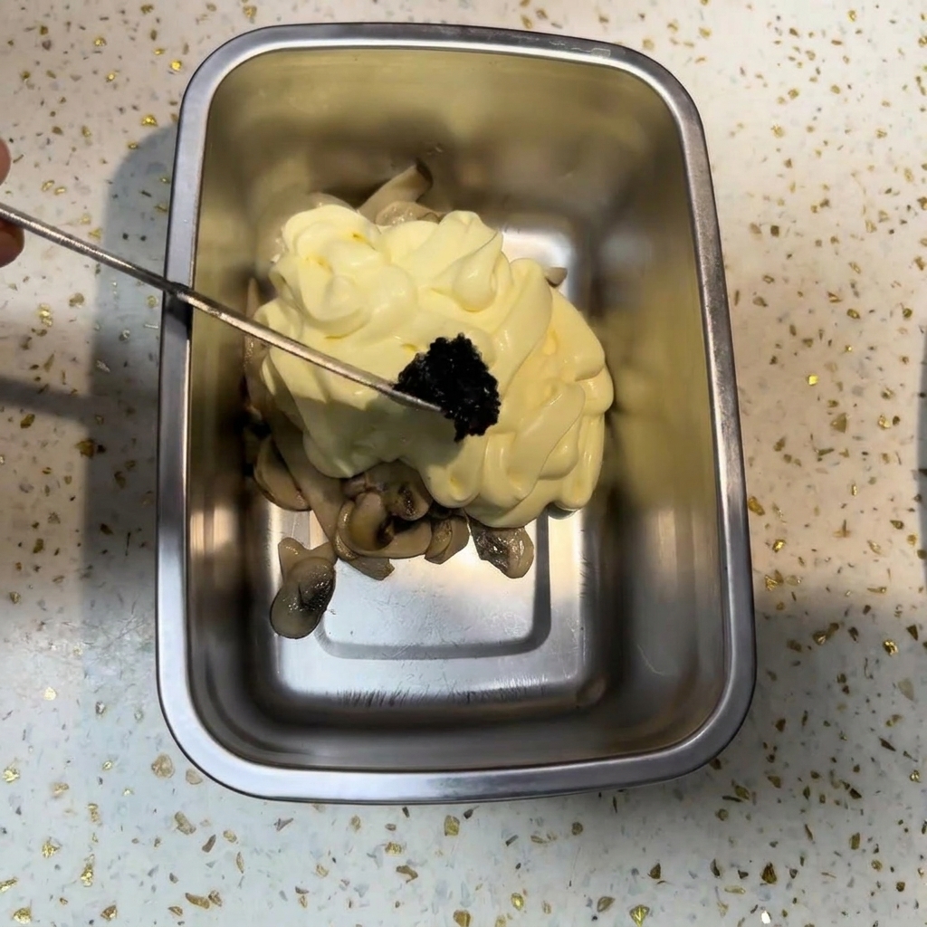 Un petit ustensile en métal déposant une portion de pâte de truffe noire sombre sur un dôme de mayonnaise et de champignons.