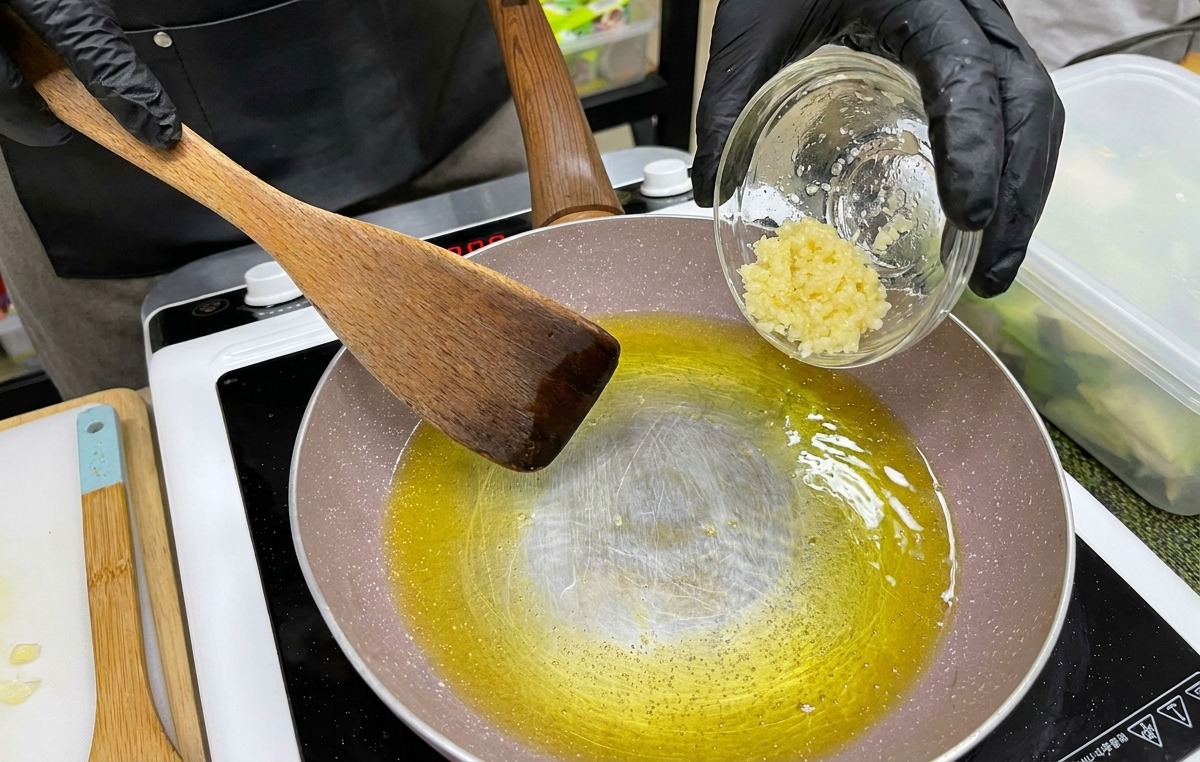 Une main avec un gant noir versant un petit bol dail émincé dans une poêle contenant du beurre fondu et de lhuile.