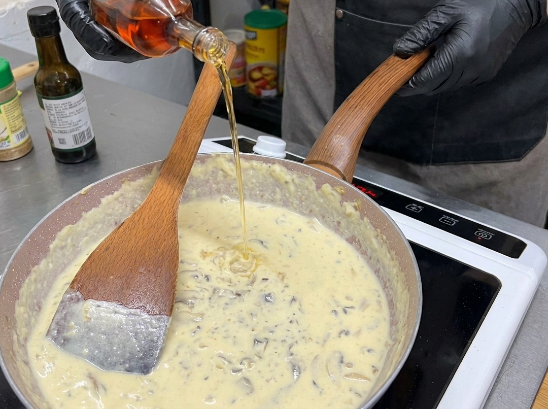 Une personne avec des gants noirs versant un liquide ambré depuis une bouteille en verre dans une poêle remplie de sauce aux champignons crémeuse.