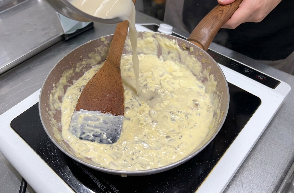 Du bouillon clair versé depuis un bol en inox dans une poêle de champignons et de crème qui mijote.