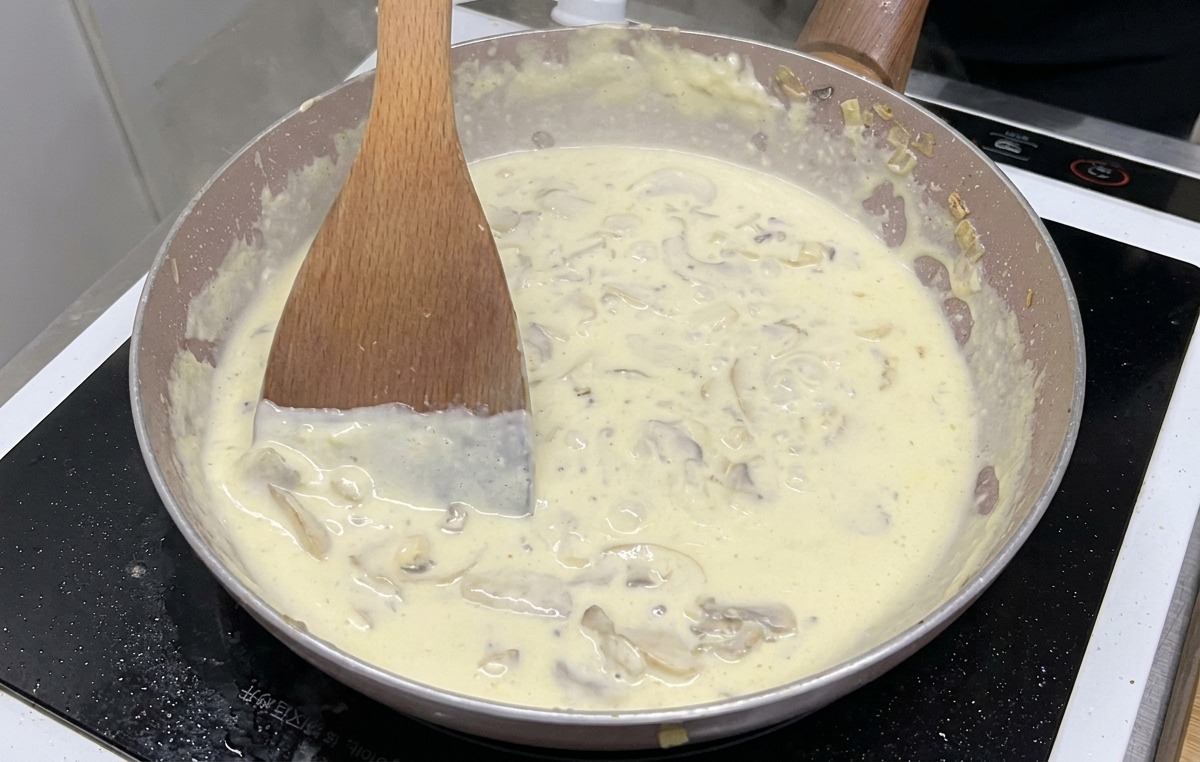 Une sauce crémeuse aux champignons avec des morceaux visibles qui bouillonne et mijote dans une poêle tout en étant remuée avec une spatule en bois.