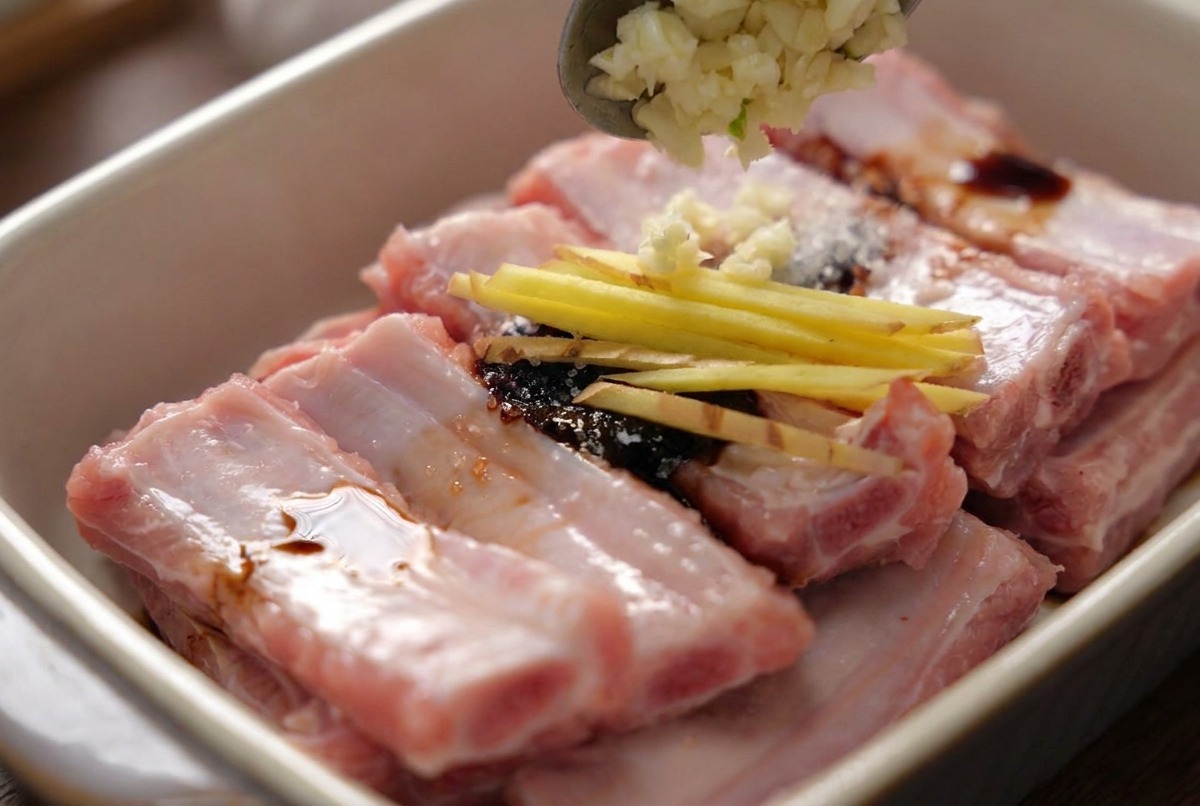 Une cuillère déposant de lail haché sur des travers de porc marinés déjà parsemés de fins bâtonnets de gingembre.