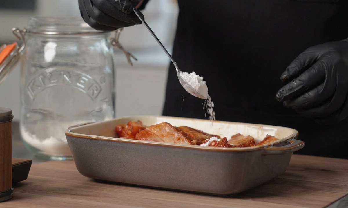 Une personne portant des gants noirs ajoute une cuillère de fécule de maïs blanche dans un plat rectangulaire rempli de travers de porc marinés.