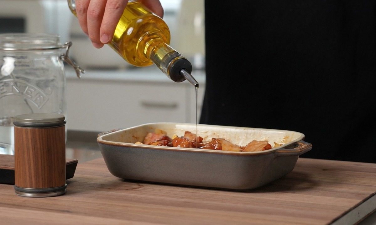 De lhuile de cuisson dorée versée depuis une bouteille en verre sur des travers de porc assaisonnés dans un plat en céramique gris.
