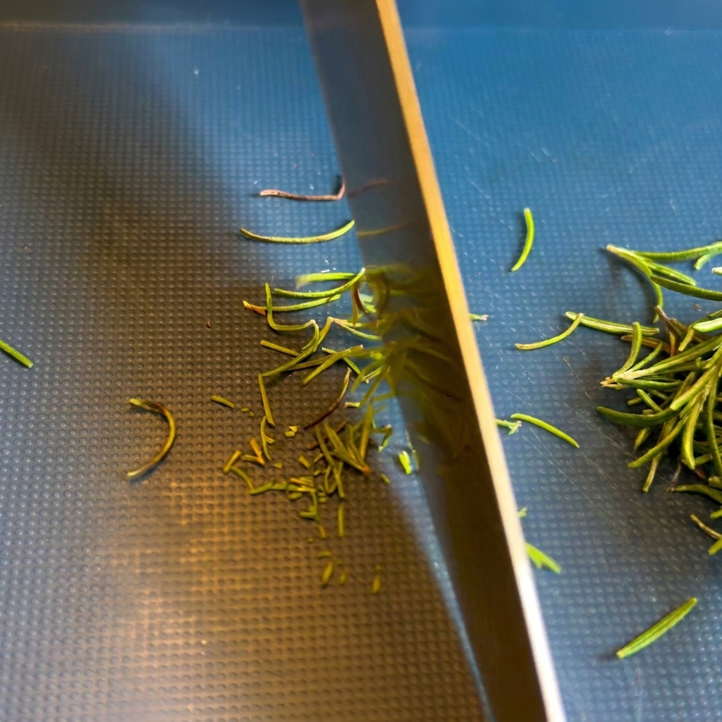 Gros plan sur la lame dun couteau de chef qui émince des aiguilles vertes de romarin frais sur une planche à découper sombre.