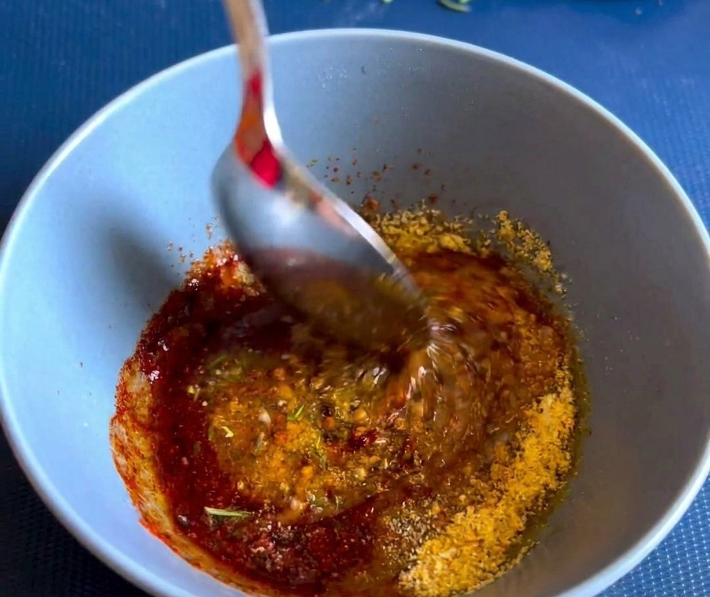Une cuillère en métal mélange une pâte dépices et dherbes épaisse et dun rouge-brun vif dans un petit bol en céramique bleu.