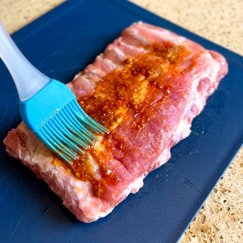 Un pinceau en silicone bleu étale une épaisse marinade rouge granuleuse sur toute la surface de ribs de porc crus posés sur une planche bleue.