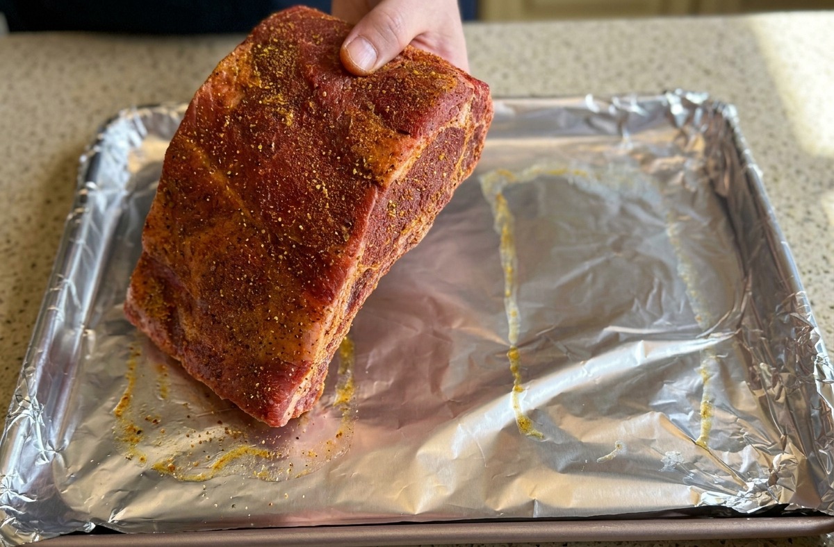 Une main dépose de grands ribs de porc bien assaisonnés sur une plaque de cuisson recouverte de papier aluminium froissé.