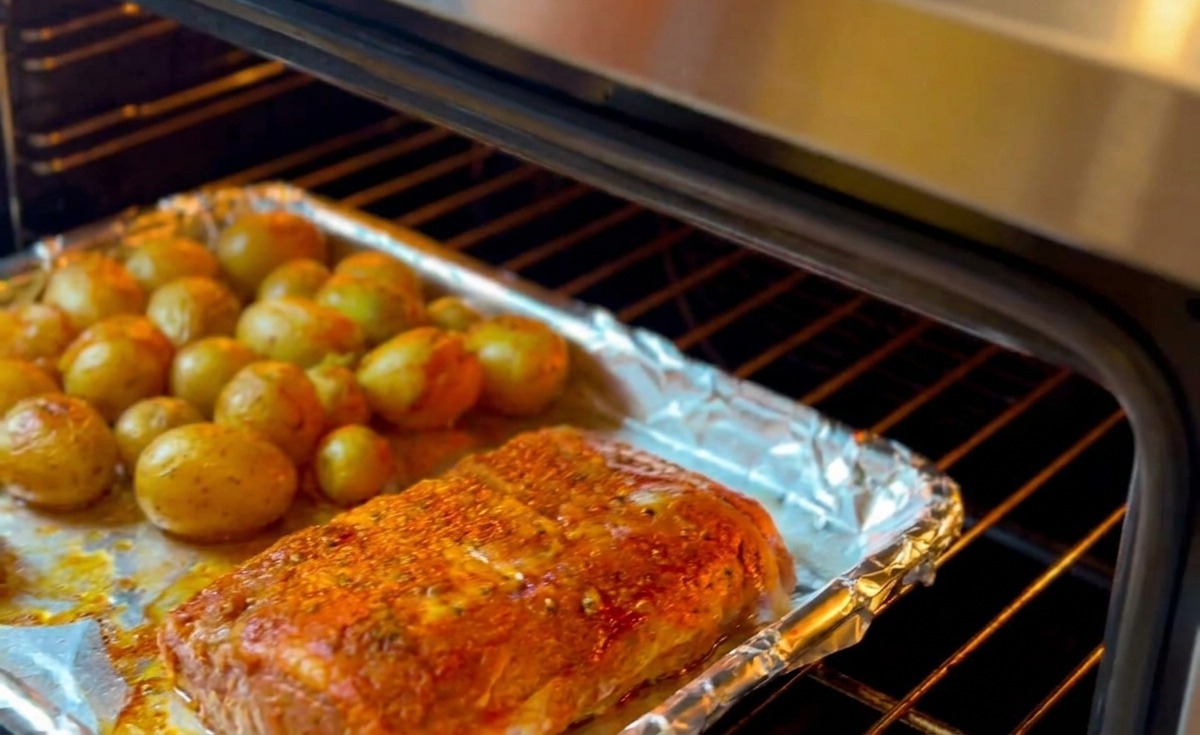 Une plaque de cuisson garnie de ribs frottés aux herbes et de petites pommes de terre jaunes est glissée dans un four préchauffé.