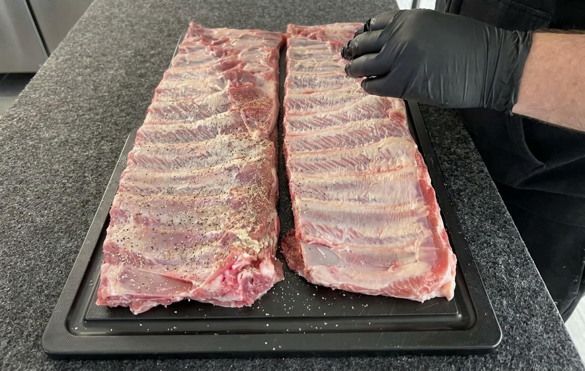 Deux morceaux de travers de porc crus assaisonnés dun mélange sec de sel, poivre et ail en poudre sur une planche à découper noire.