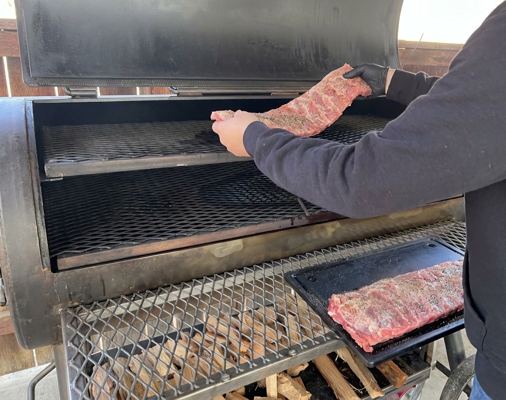 Une personne portant un sweat à capuche noir et des gants noirs déposant un morceau de ribs assaisonné sur la grille supérieure dun grand fumoir offset.