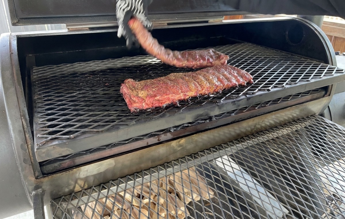Une main dotée dun gant anti-chaleur à motifs retournant un morceau de ribs partiellement fumé sur la grille métallique dun fumoir.