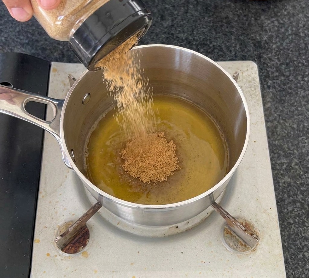Une main versant de la cassonade depuis un bocal transparent dans une casserole en inox contenant du vinaigre de cidre sur un réchaud.