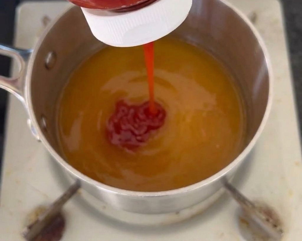 Un filet de ketchup rouge versé depuis un flacon souple dans une casserole contenant une base de sauce liquide jaune.