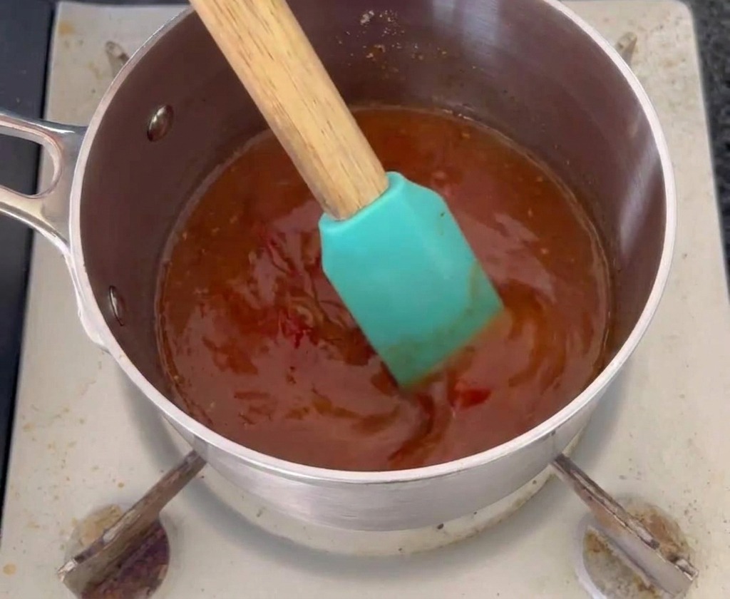 Une spatule en silicone bleu canard remuant une sauce mop orange-rouge foncé dans une petite casserole en acier inoxydable.