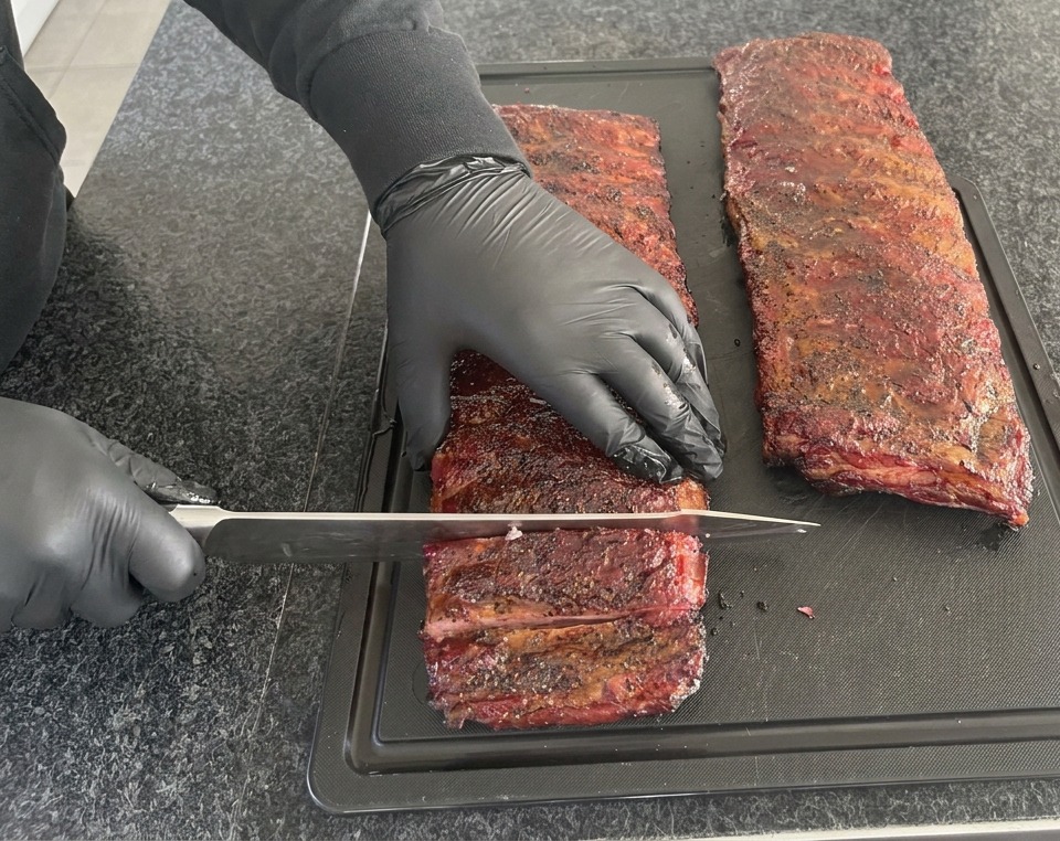 Une personne portant des gants en nitrile noirs utilisant un grand couteau de chef pour découper un long morceau de ribs fumé rouge foncé sur une planche noire.