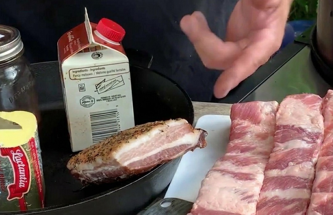 Plusieurs longues bandes de travers de porc crues étalées sur une planche à découper blanche, à côté dingrédients comme du beurre et des bleuets frais.