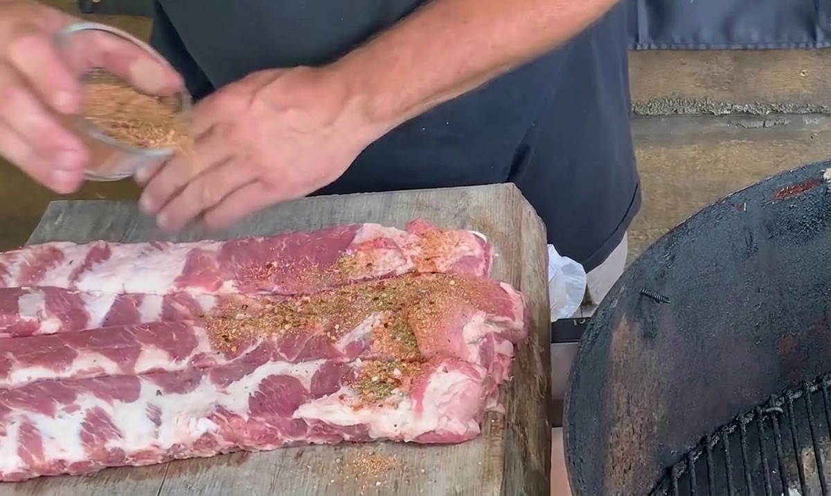 Des mains saupoudrant un mélange dépices brun-rouge sur des travers de porc crus posés sur une planche à découper en bois.