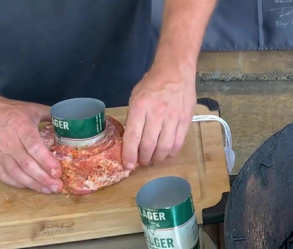 Des mains attachant soigneusement de la ficelle blanche autour dune bande de porc assaisonnée enroulée autour dune canette de bière verte.