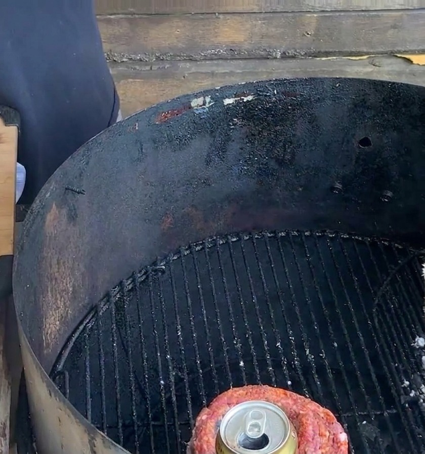 Une coupe de porc ficelée contenant une canette en métal repose sur la grille noire dun barbecue au charbon à côté dune planche de préparation.