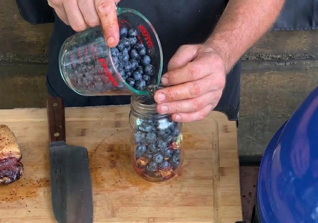 Des bleuets frais versés depuis un verre doseur dans un bocal en verre contenant déjà de lail haché et des morceaux de bacon.