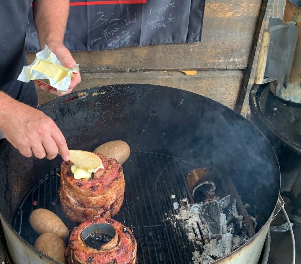 Une cuillère en bois déposant une noix de beurre sur la viande sur un barbecue au charbon, à côté de pommes de terre entières.