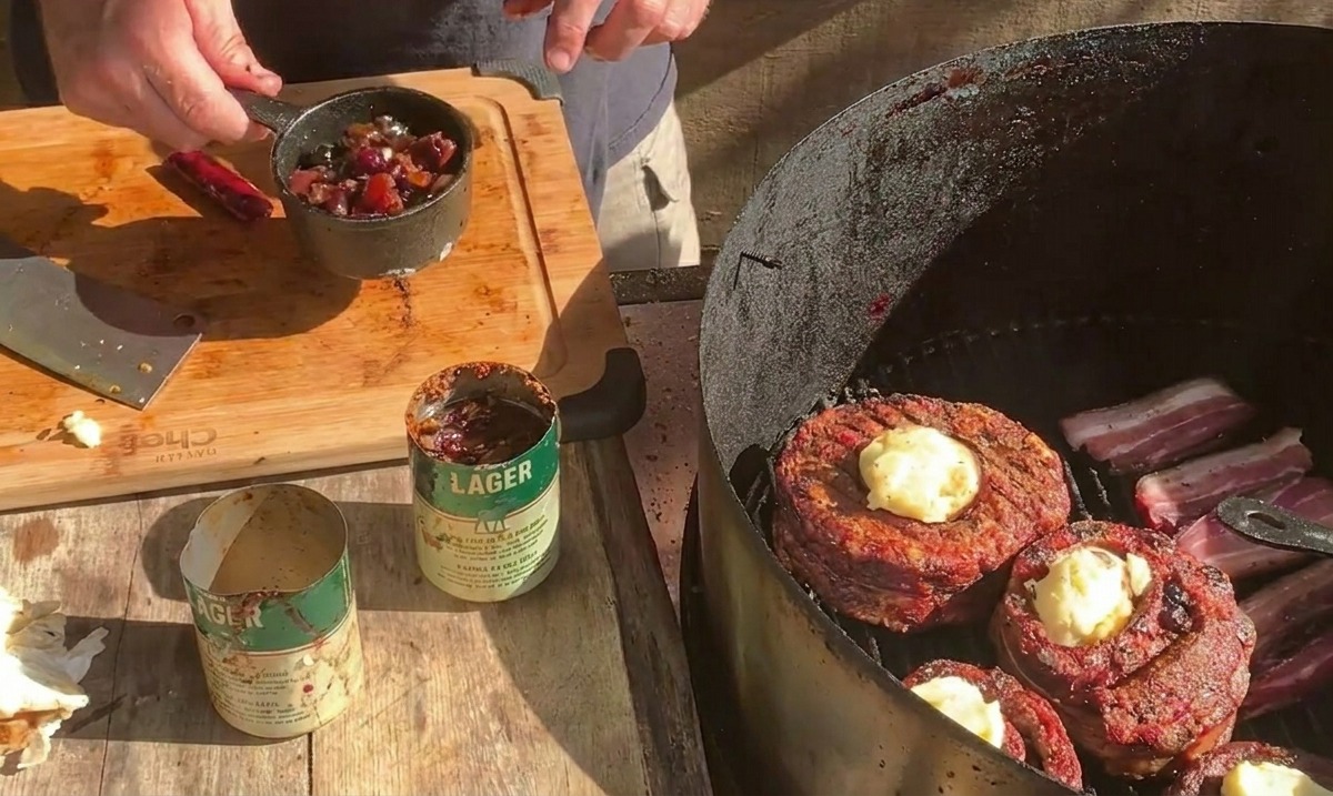 Une personne tenant une petite casserole en métal remplie de sauce aux bleuets au-dessus dun barbecue où des coupes de ribs entourées de bacon reçoivent du fromage.
