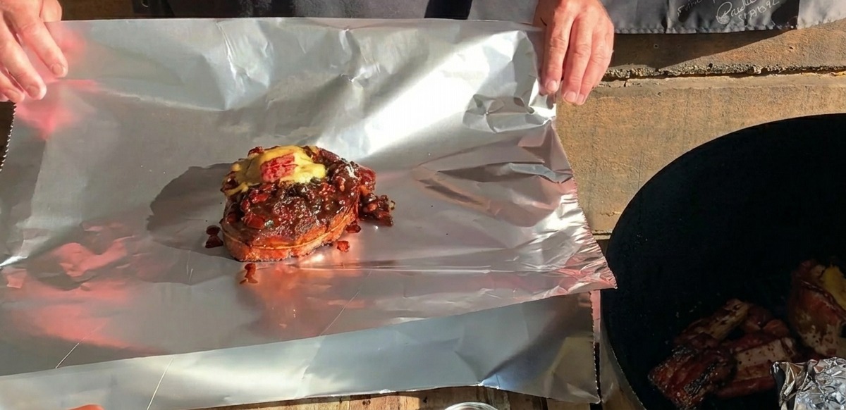 Une coupe de ribs cuite au barbecue, surmontée de fromage fondu, posée sur une grande feuille de papier daluminium prête à être enveloppée.