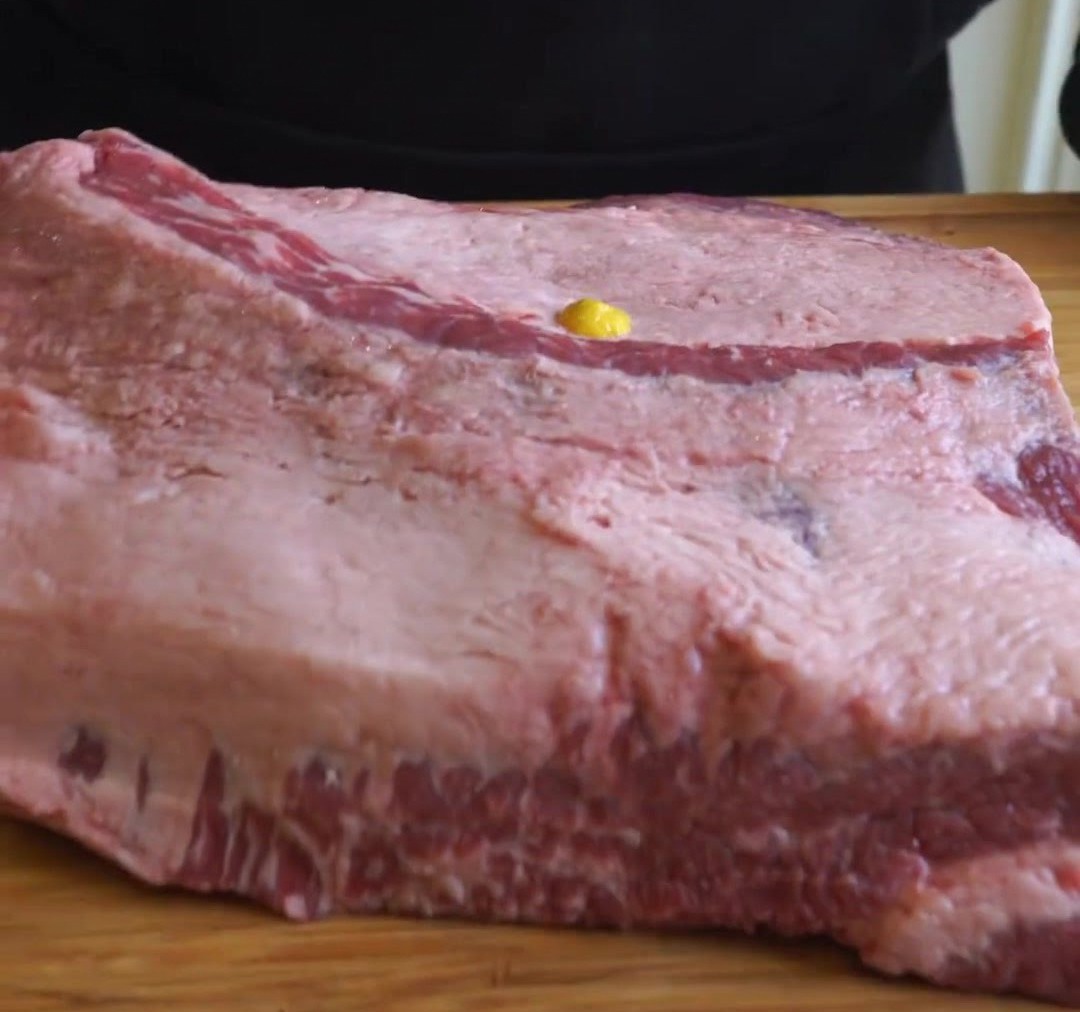 Un filet de moutarde jaune versé sur une grande poitrine de bœuf crue posée sur une planche en bois.
