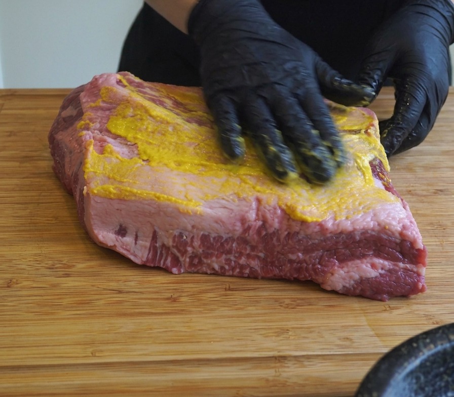 Des mains gantées de noir étalant de la moutarde jaune et du sel sur un grand brisket posé sur une planche à découper.