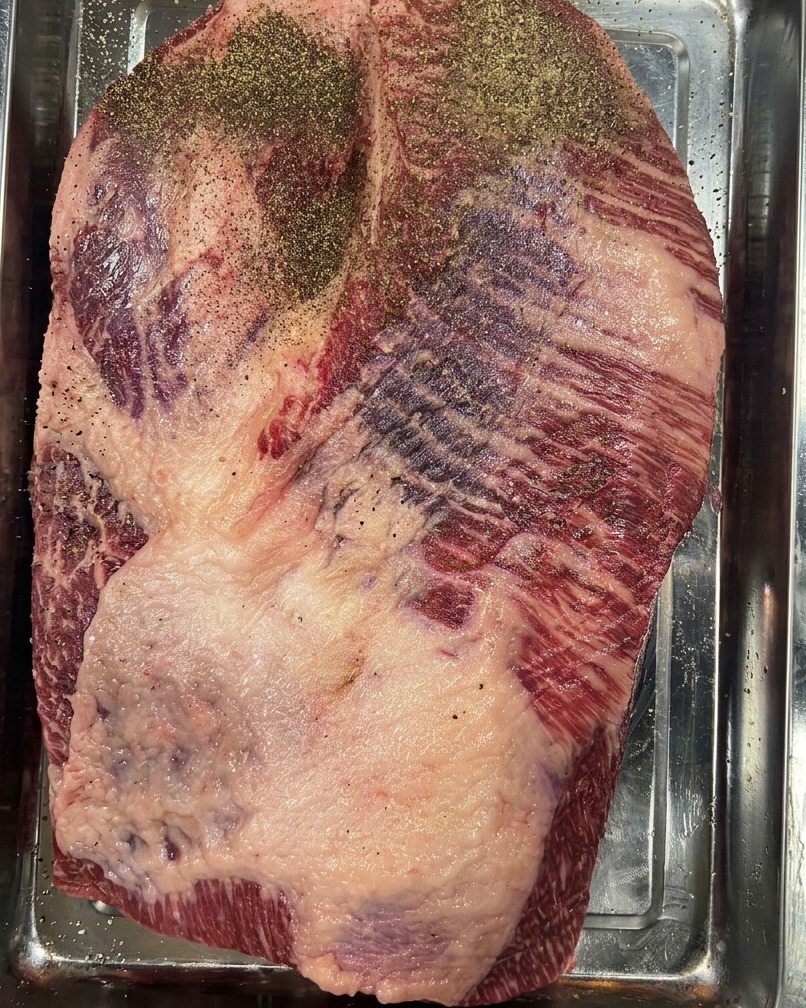 Une poitrine de bœuf crue dans un plat en inox, recouverte dune épaisse couche de poivre noir moulu.
