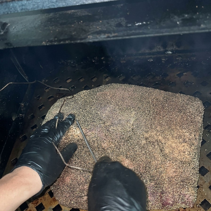 Une personne en gants noirs insérant la sonde métallique dun thermomètre dans un brisket assaisonné au sein dun fumoir sombre.