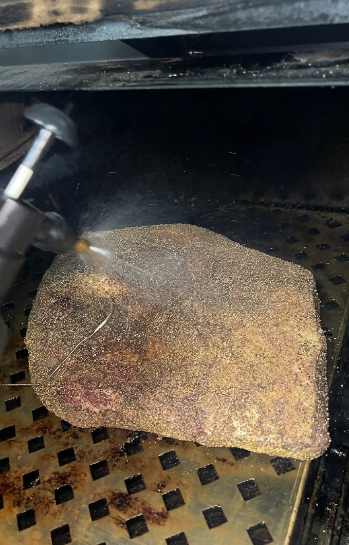 Une fine brume de vinaigre de cidre vaporisée sur un brisket en train de développer une croûte sombre dans le fumoir.