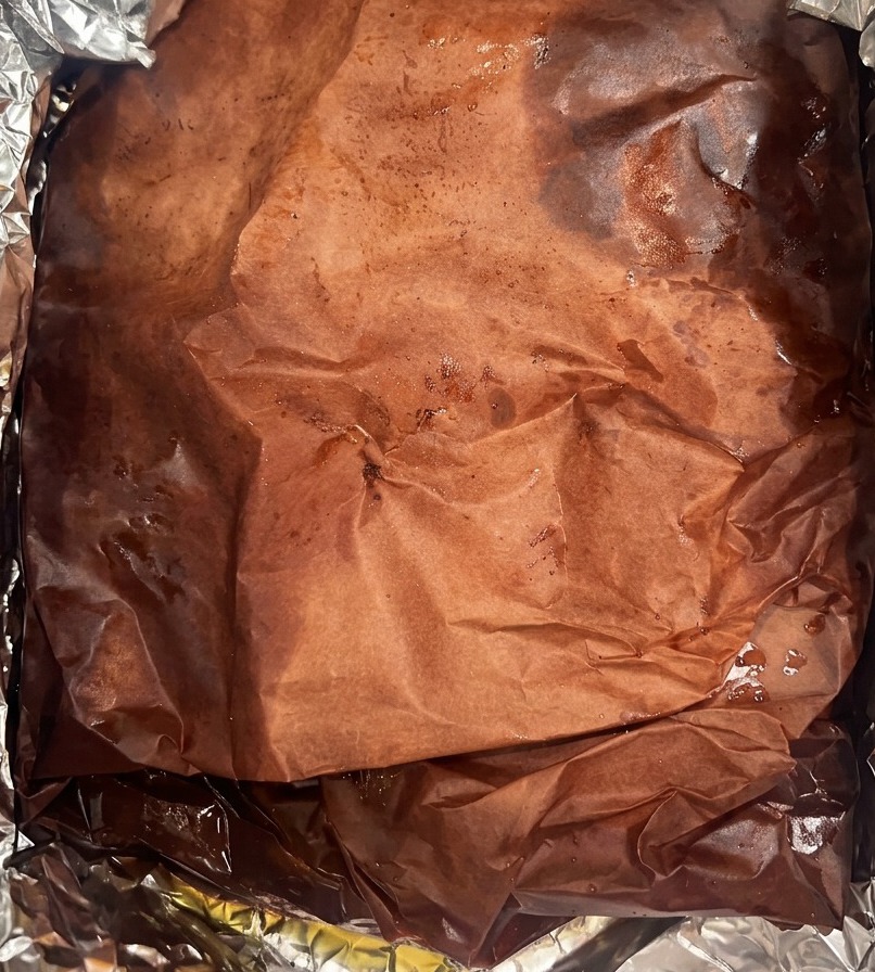 Un grand brisket enveloppé dans du papier boucher sombre et imprégné de jus, déposé dans un bac doublé daluminium.