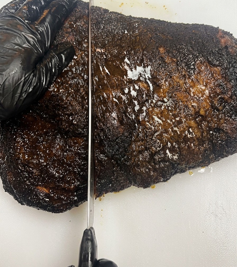 Une main gantée de noir maintenant un brisket fumé à la croûte sombre sur une planche à découper tandis quun long couteau tranche la viande.