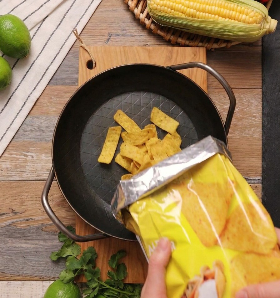 Des chips de maïs triangulaires jaunes versées depuis un paquet jaune dans une poêle en fonte noire posée sur une surface en bois.