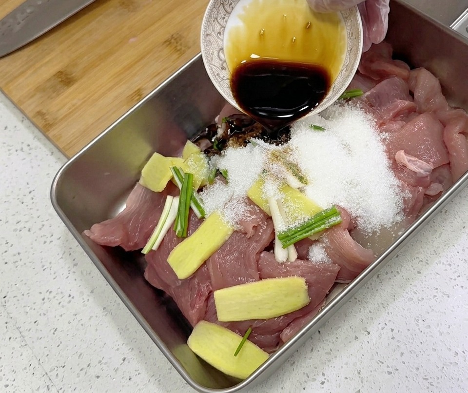 Un assaisonnement liquide sombre est versé dun petit bol finement décoré sur des tranches de porc cru, du gingembre et des oignons verts dans un plateau métallique.