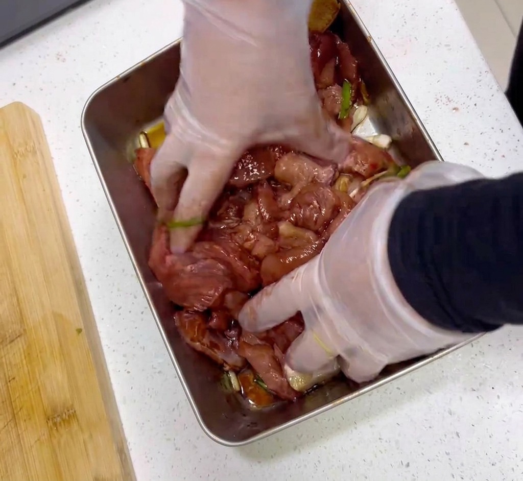 Des mains portant des gants en plastique transparent mélangent des tranches de viande crue avec une marinade rouge foncé dans un plateau en acier inoxydable.