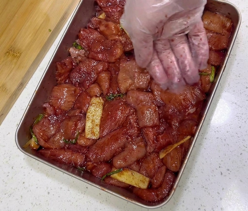 Des tranches de porc rouge marinées dans un plateau en acier inoxydable sont assaisonnées avec une cuillerée de gros flocons de piment rouge.