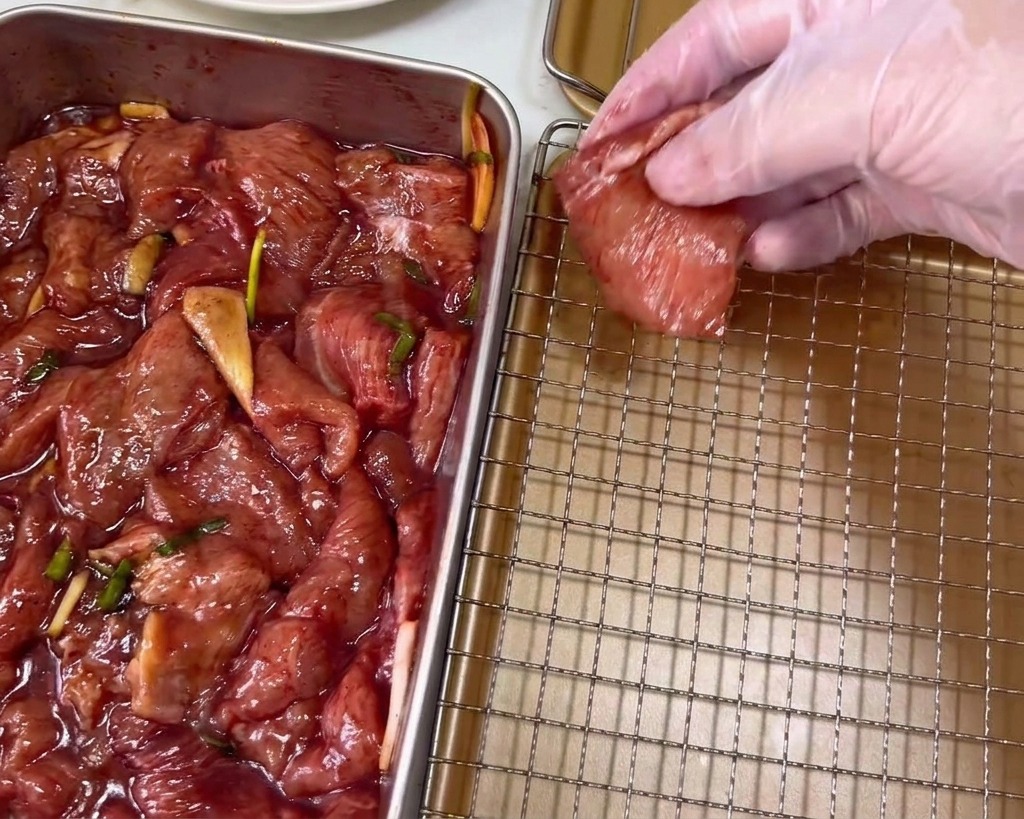 Une main dans un gant en plastique transparent dépose une tranche de porc mariné rouge foncé sur une grille de refroidissement dorée.