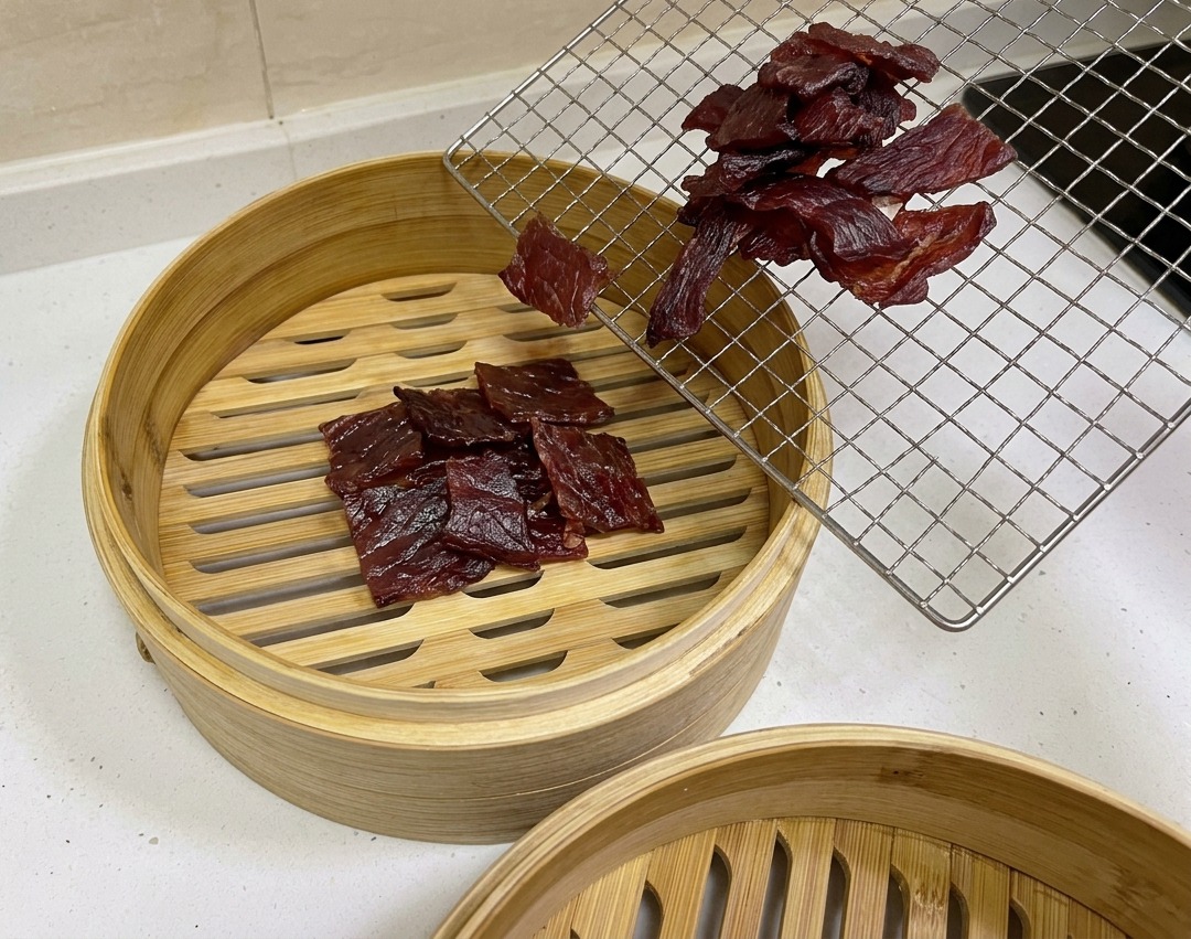 Des morceaux de porc séché à lair brun rougeâtre foncé sont déplacés dune grille de refroidissement métallique vers un panier en bambou circulaire pour cuisson à la vapeur.