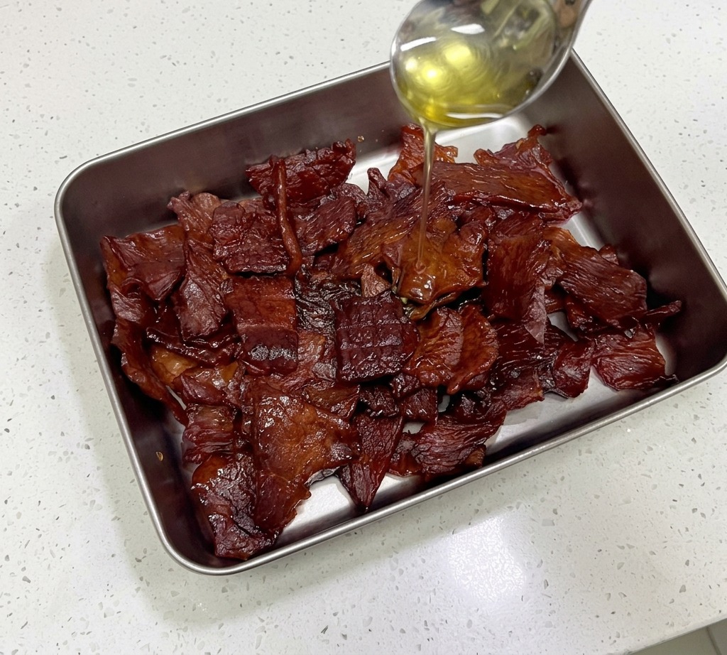 Du miel doré est versé dune cuillère en acier inoxydable sur un monticule de lanières de viande de porc dun rouge acajou brillant dans un récipient en métal.