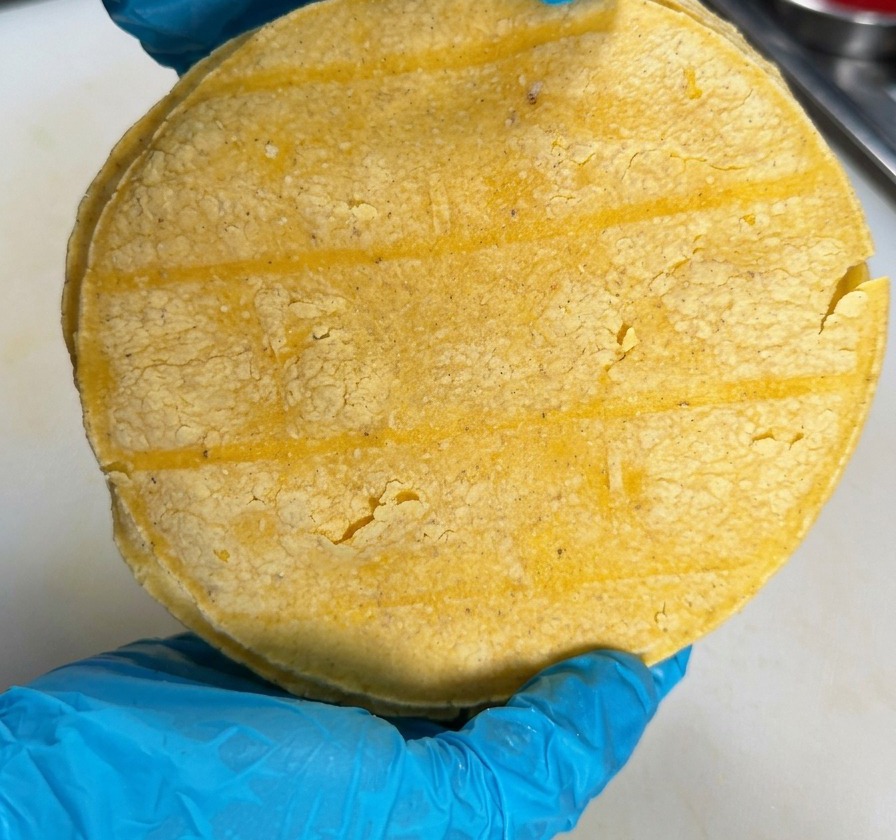 Une personne portant des gants bleus tient une pile de tortillas de maïs jaune au-dessus dun plan de travail blanc.
