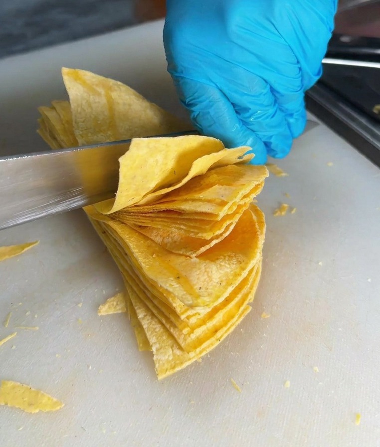 Un couteau de chef tranchant découpe une pile de tortillas de maïs en triangles sur une planche à découper blanche.