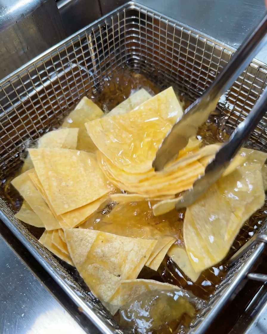 Des pinces en métal déposent des morceaux de tortillas en forme de triangle dans le panier en grillage dune friteuse.