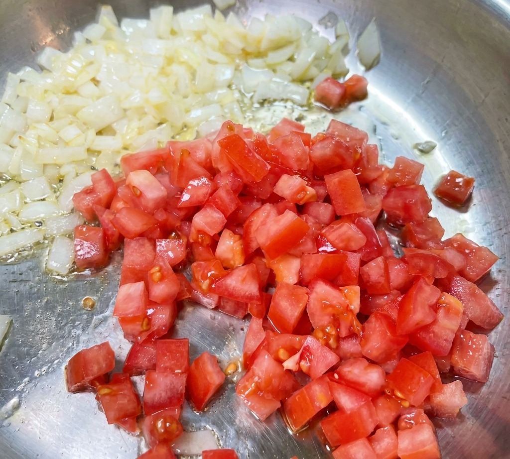 Des dés de tomates rouges mélangés à des oignons revenus et translucides dans une poêle.