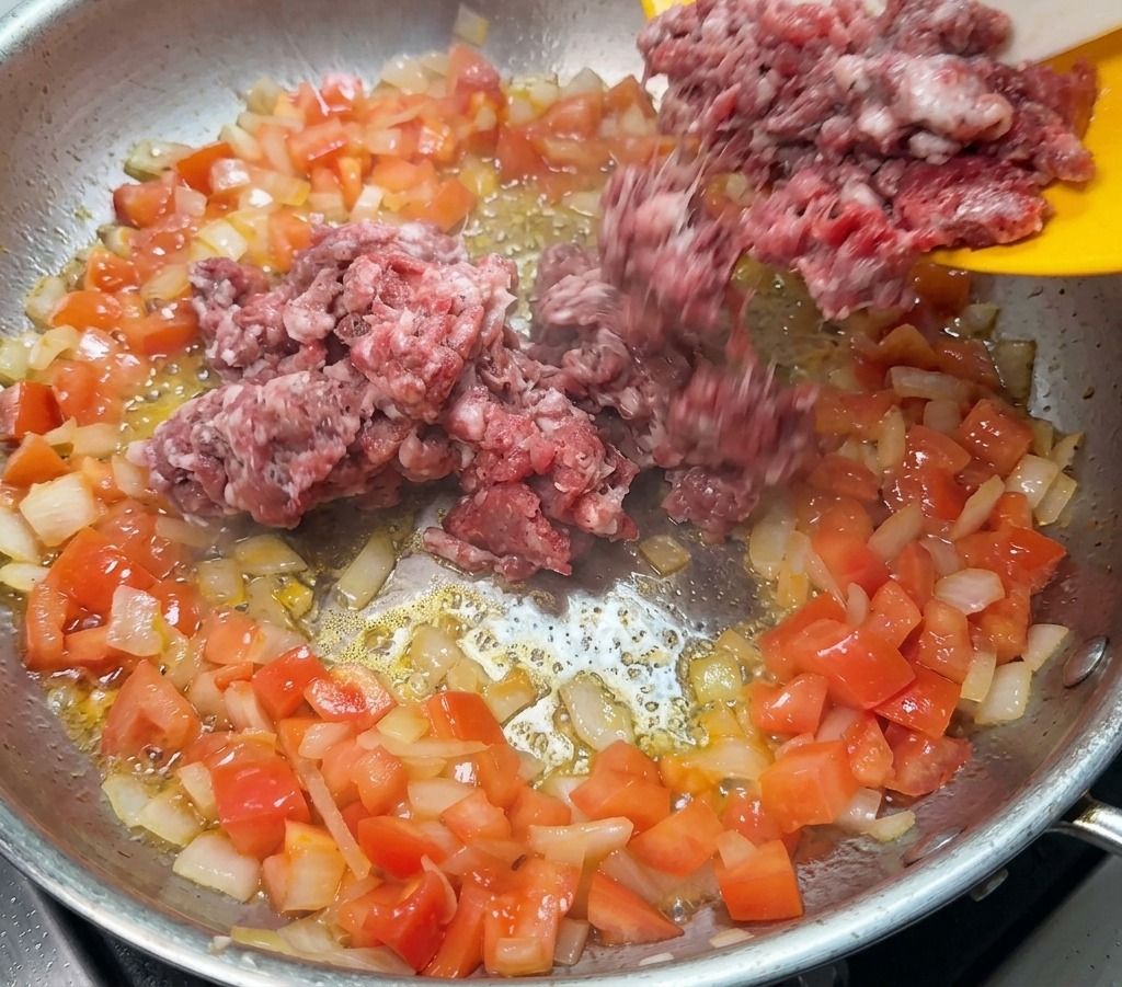 De la viande hachée crue ajoutée depuis une assiette jaune dans une poêle contenant des oignons et des tomates.