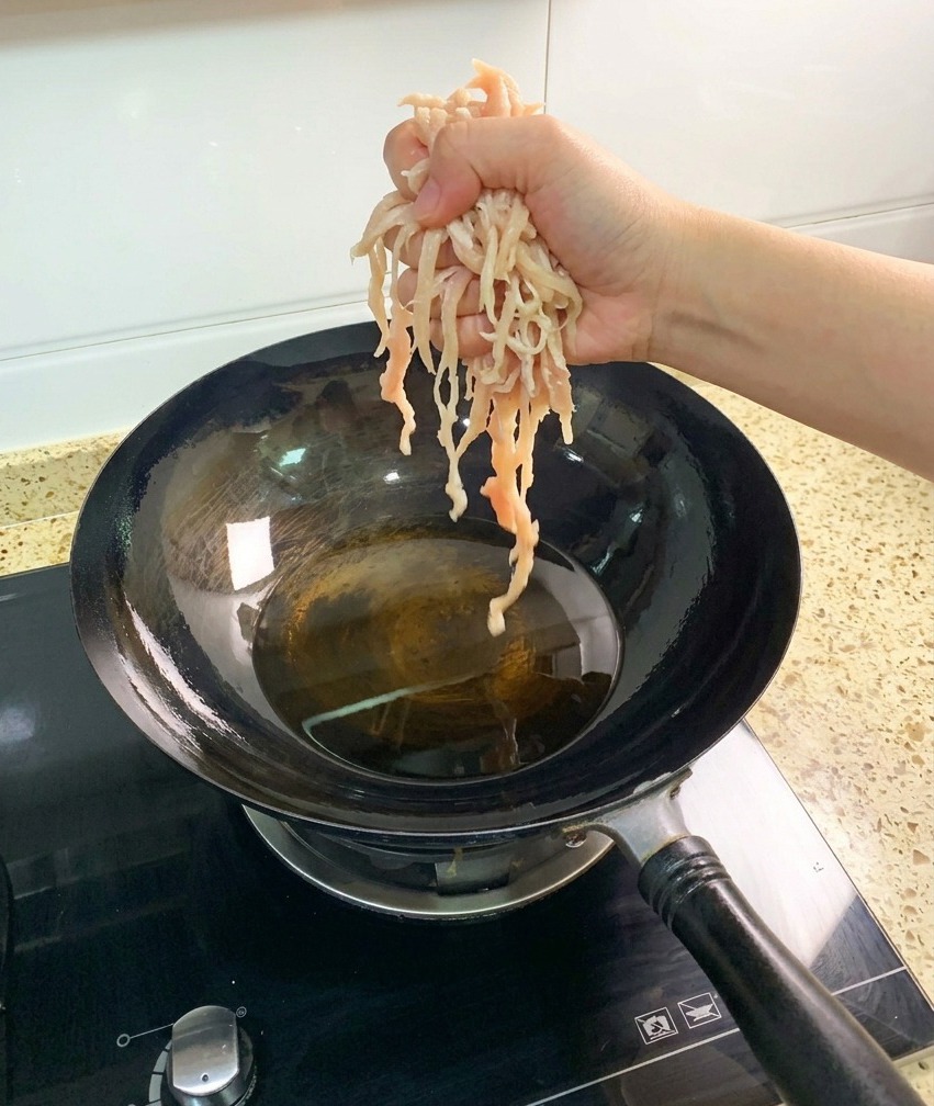 Une main déposant des morceaux pâles de blanc de poulet effiloché cru dans un wok sombre rempli dhuile de cuisson chaude.