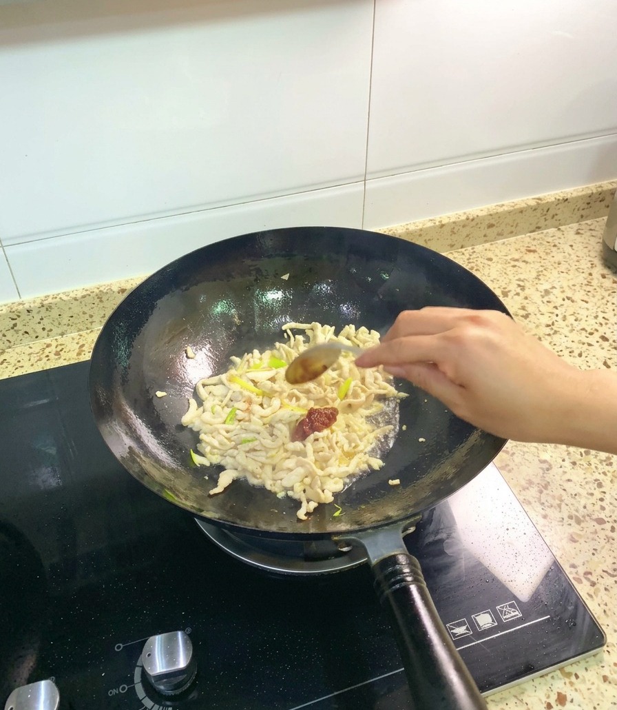 Une cuillère en acier inoxydable ajoutant une généreuse portion de pâte de haricots rouges de Pixian dans un wok de poulet sauté et daromates.