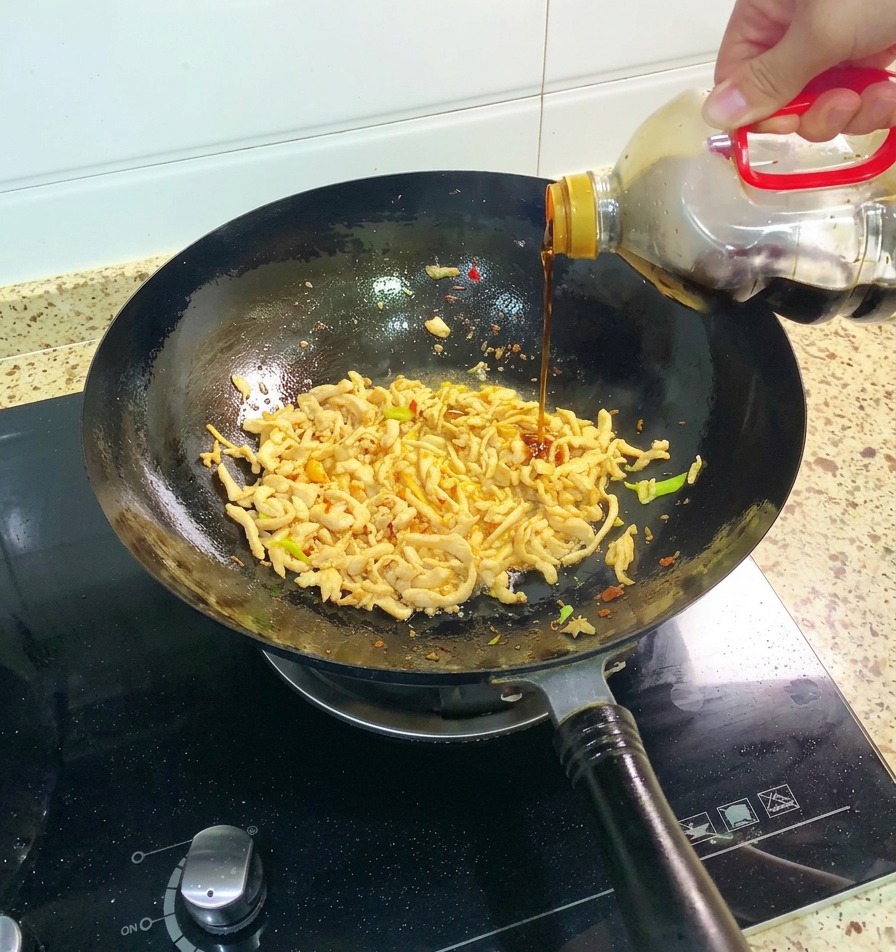 Une main versant de la sauce soja foncée depuis une grande bouteille dans un wok contenant du poulet effiloché et des aromates verts.